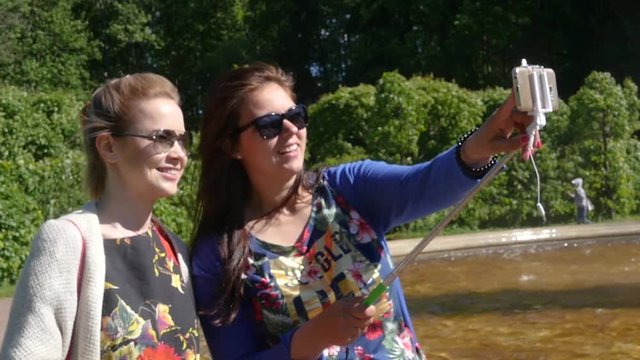 Two young girlfriends doing photo on phone and selfie stick in green park. slow motion. 1920x1080. full hd
