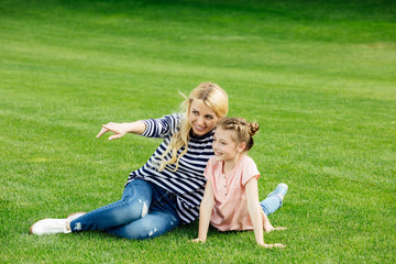 beautiful happy mother and daughter pointing away with finger while sitting on green meadow