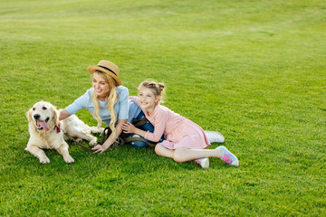 happy beautiful mother and daughter with golden retriever dog sitting on green lawn at park
