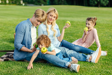 Fototapeta premium happy young family with two kids eating green apples while resting on grass in park