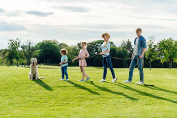 Naklejka premium side view of happy young family with pet walking on green meadow at park