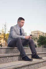 Obraz premium one young Caucasian man smiling, business suit, formal wear, ordinary common person portrait, outdoors sitting on stairs steps, using looking smart phone