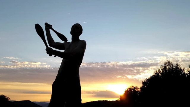 Silhouette of a lean sporty young male exercising outdoor with Indian clubs, with beautiful sunset in the background