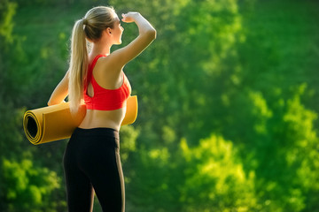 A young blonde in a red top and black pants holds a mat for a fountain in the nature. A woman prepares for gymnastics