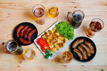 Mugs of beer and glasses of beer, plate of roasted potato and frying pans with  grilled sausages