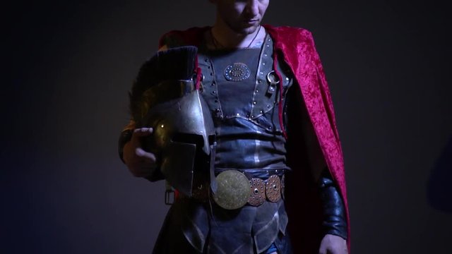 A serious gladiator stands in leather armor and a raincoat and in his hand holds a metal helmet and looks into the camera, bit slow motion