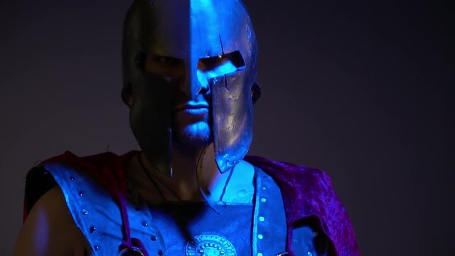 The Roman gladiator in a heavy helmet, leather armor and red raincoat looks into the camera standing in the ultraviolet light, slow motion shooting