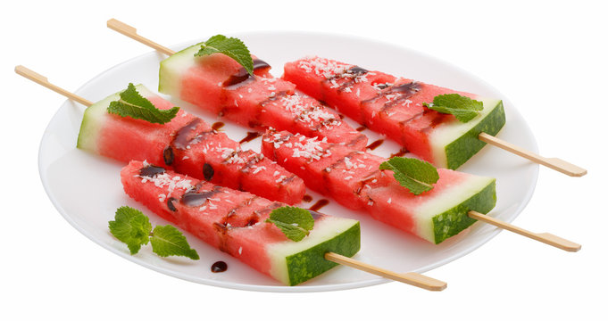 Slices of watermelon with stick on white plate isolated on white background. It looks like ice cream. Decorated with mint leaves, coconut and chocolate syrup.