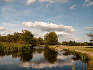 long cloud and blue sky reflected in river with trees in the country lady walking