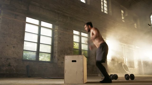 Athletic Shirtless Fit Man Energetically Box Jumps in Hardcore Gym doing Part of Cross Fitness Training Program. Man is Sweaty from Intense Workout/ Exercise, Gym is in Industrial Factory Location.