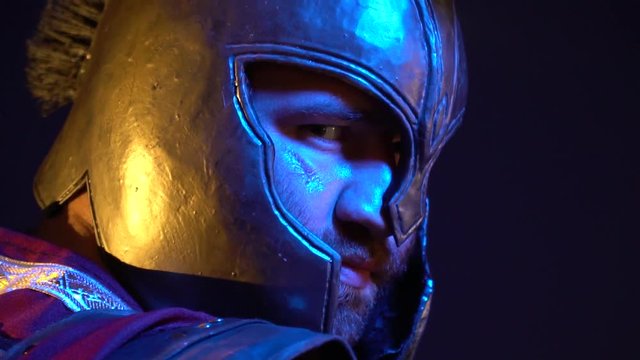 Unshaven Roman gladiator with a scar on his face in a helmet and armor stands with his back, looks around and turns on the camera