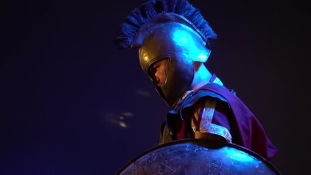 The Roman gladiator in leather armor and heavy helmet holds a shield and stands with a thoughtful look