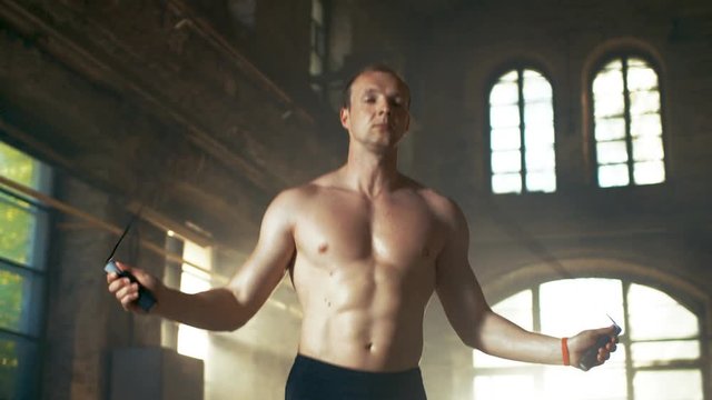 Athletic Shirtless Fit Man Exercises with Jump / Skipping Rope in a Deserted Factory Hardcore Gym. He's Covered in Sweat from His Intense Cross Fitness Training