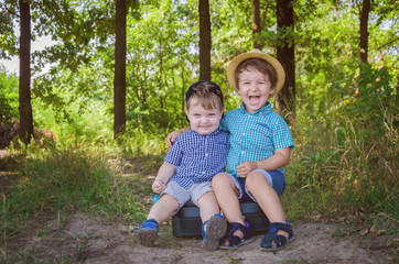 Two little boys are sitting on a suitcase in the woods on the path