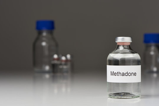  A ampoule of methadone (English label) stands on white surface against gray background on the right. Further laboratory bottles can be seen in the background. (English label) in cross-format