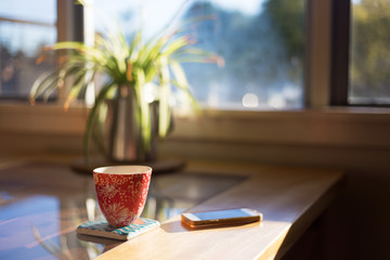 A cup of coffee on a coaster next to smart phone on table with bright morning window light...