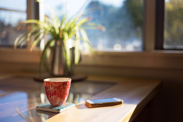  A cup of coffee on a coaster next to smart phone on table with bright morning window light streaming through.
