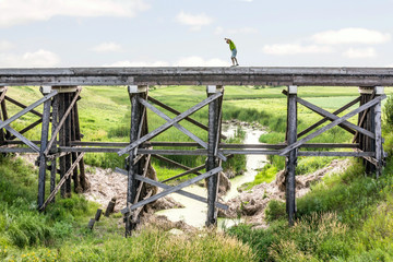 horizontal image of a caucasian man taking a risk and walking on the edge of a very tall  old abandoned railroad track with a stream flowing underneath with green vegetation in the summer time.