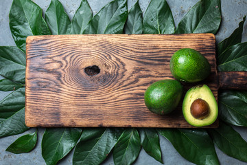 food background with fresh avocado, avocado tree leaves and wooden cutting board. Harvest concept, Guacamole ingredients. Healthy fat, omega 3. Half of avocado. Top view. Copy space.