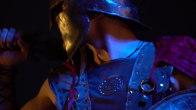 The Roman gladiator in a beautiful armor and raincoat stands with a shield and sword on his shoulder in a spectacular pose and looks the side