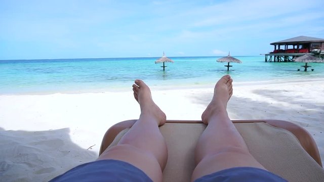 Tourist legs bare feet sitting on golden sand beach bench point out to tropical ocean paradise