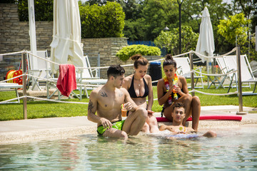 Young handsome man and attractive women in swimwear sitting at pool relaxing under sun. 