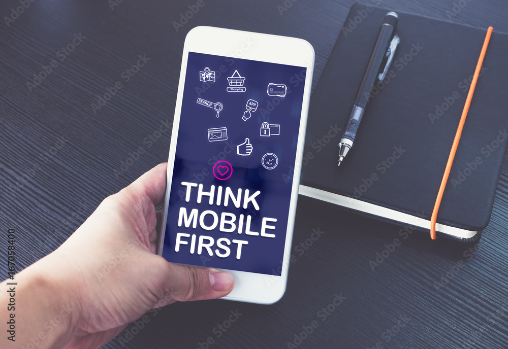 Aufkleber Hand holding mobile phone with Think mobile first word and feature icon over notebook on black wood table background,Digital marketing concept ,office desk