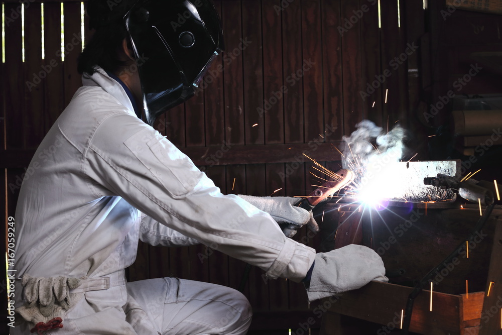 Wall mural side view of professional welder in white uniform with protective helmet welding steel with spark in