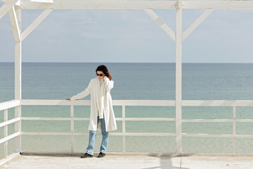 Young woman (brunette) in a white coat on the promenade near the sea.