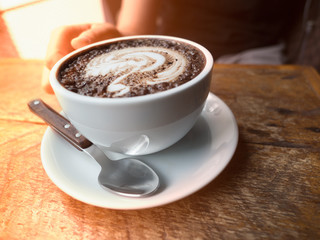 Hot chocolate latte art milk cup in woman hand over wooden table with sunrise warm light background in a morning at coffee shop