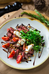 On a plate, on a wooden surface, close-up roast beef salad with rocket salad, cherry tomatoes, mozzarella, balsamic sauce, rosemary, spices, nuts, tall kitchen, restaurant, trendy delicious food