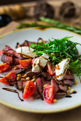 On a plate, on a wooden surface, close-up roast beef salad with rocket salad, cherry tomatoes, mozzarella, balsamic sauce, rosemary, spices, nuts, tall kitchen, restaurant, trendy delicious food