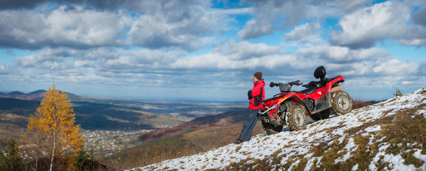 Panorama. Man standing near ATV quad bike, looking into the distance on snowy mountain top. On the background blue cloudy sky, mountains and the town in the valley with copy space. Autumn © anatoliy_gleb