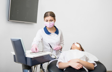 Woman dentist with female patient before treatment in the modern dental office. Doctor picking up dental tools, wearing white uniform, gloves, mask,. Dentistry. Stomatology equipment