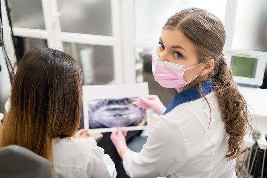 Female young dentist examining x-ray image with female patient in dental office, looking to the camera and preparing for treatment. Dentistry