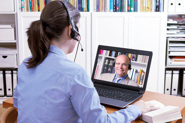 Woman with a headset in front of her laptop and a book making an online video call with her...