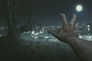 Obraz na płótnie Canvas Halloween Concept, The point of view of the zombie's right hand, with the background of an old tree and a city lit by the light of the full moon.