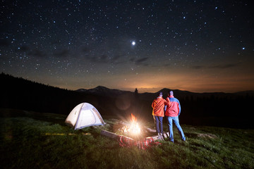 Fototapeta premium Night camping in the mountains. Rear view of romantic couple tourists standing at a campfire near illuminated tent under starry sky at night. Low light