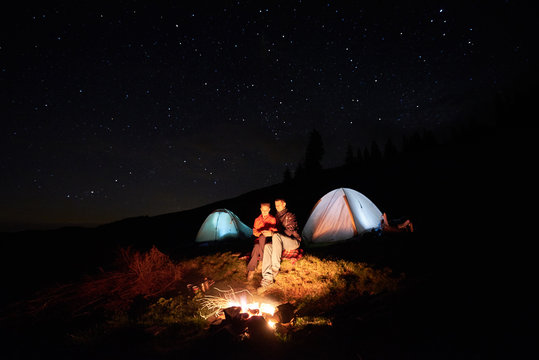 Night camping in the mountains. Couple tourists sitting at a campfire near two illuminated tents under night starry sky. Long exposure