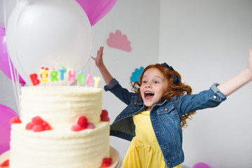 cute happy red haired girl with raised hands jumping near birthday cake