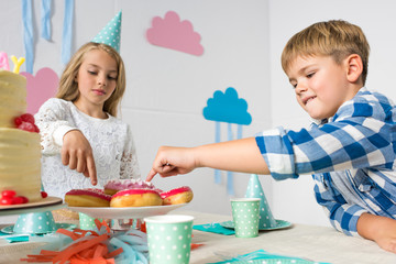 cute little children pointing with fingers at tasty doughnuts while sitting at birthday table