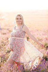 Fototapeta na wymiar Smiling pregnant woman wearing stylish dress with floral pattern in meadow. Looking at camera. Motherhood. Maternity.