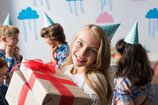 adorable smiling girl holding birthday presents while friends having fun behind