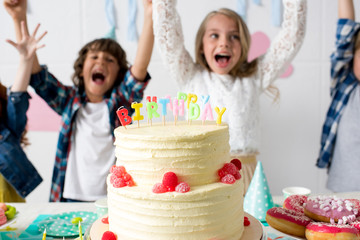 close-up view of delicious birthday cake on festive table and cheerful kids raising hands behind