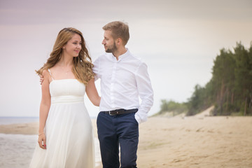 Young happy just married couple walking holding hands on the beach along the sea on summer evening