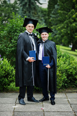 Two intelligent and handsome graduates in graduation gowns and caps posing with their diplomas.