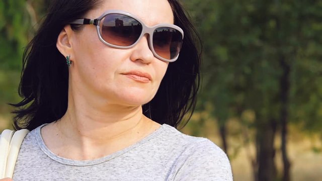 A woman in sunglasses stands against the background of green trees.