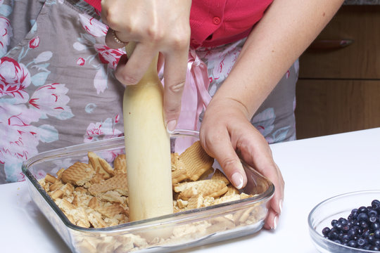 A woman is crumbling a biscuit cookie. Delicate cheesecake with blueberries. Ingredients: cookies and blueberries stand in containers on the table.