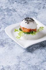 Mini rice sushi burger with smoked salmon, green salad and sauces, black sesame served on white square plate over gray concrete background. Modern healthy food