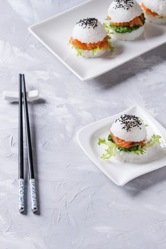 Mini rice sushi burgers with smoked salmon, green salad and sauces, black sesame served on white square plate with chopsticks over gray concrete background. Modern healthy food